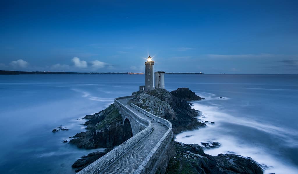 Bild mit einem Leuchtturm auf einer Felsenklippe im weiten Ozean. Das Licht leuchtet.